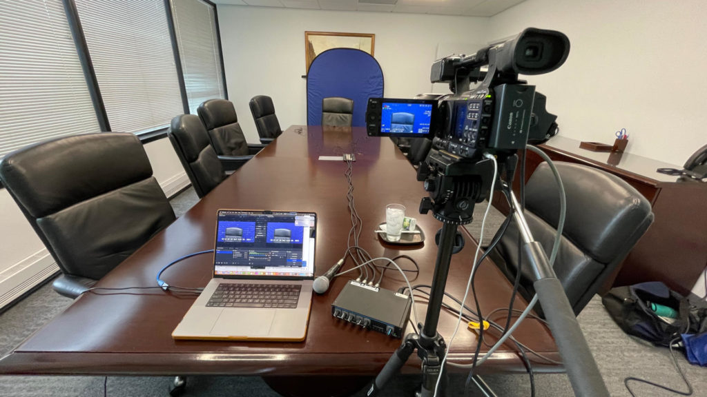 Legal videographer equipment essentials. Pictured is a high definition camera, a 4 channel audio mixer, a capture card, a laptop, 4 microphones and xlr cables and a flexi-backdrop.